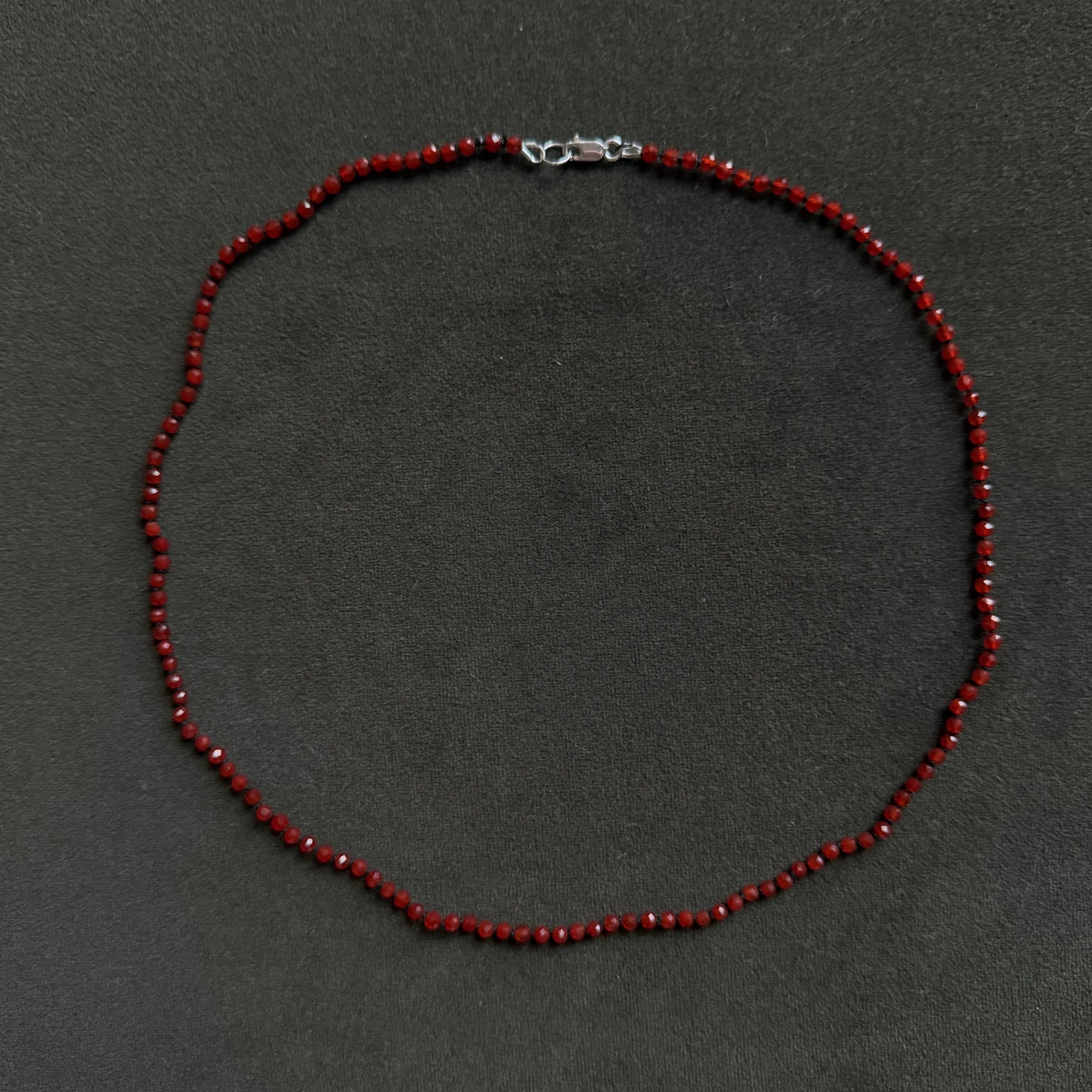 A beaded necklace made of small dark red faceted beads with a silver lobster clasp, laid flat on a dark gray textured background.