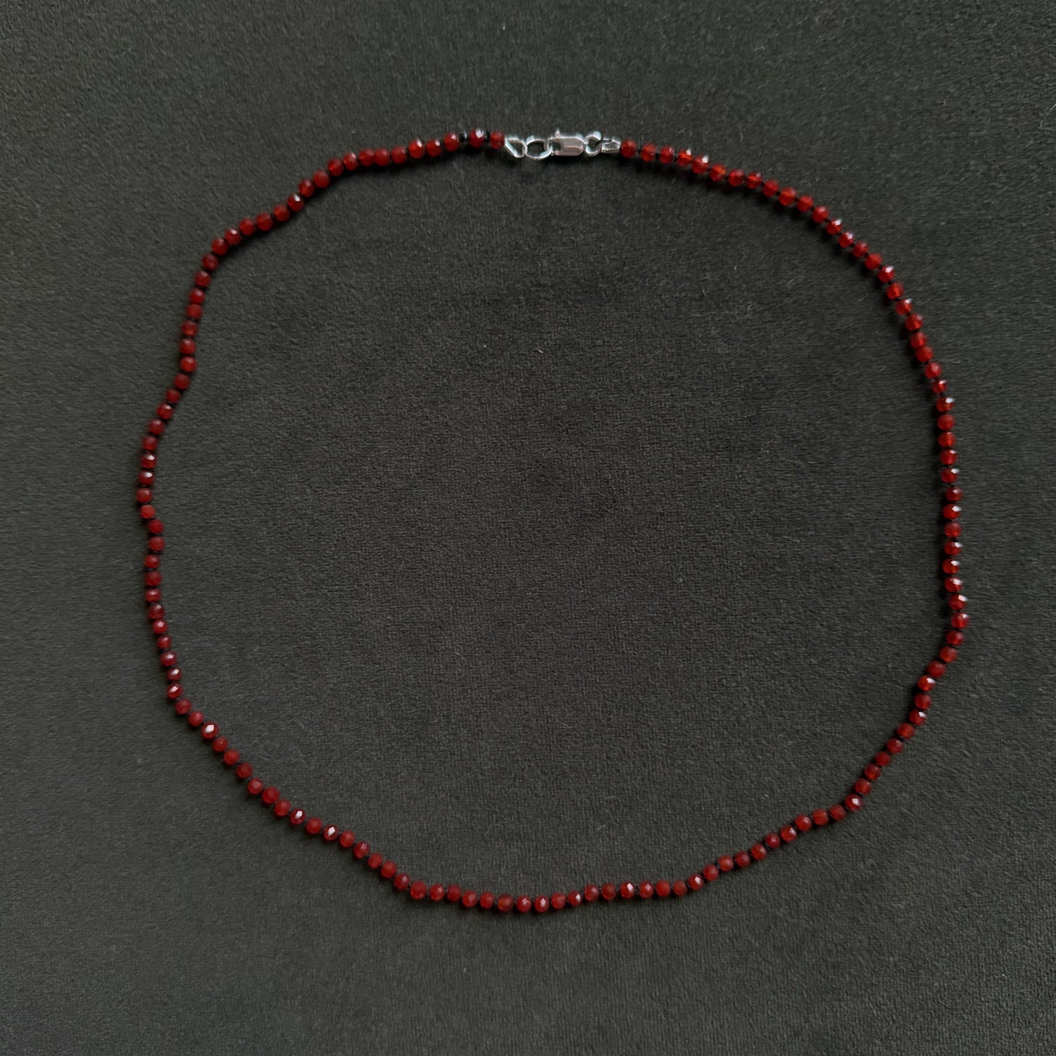 A beaded necklace made of small dark red faceted beads with a silver lobster clasp, laid flat on a dark gray textured background.