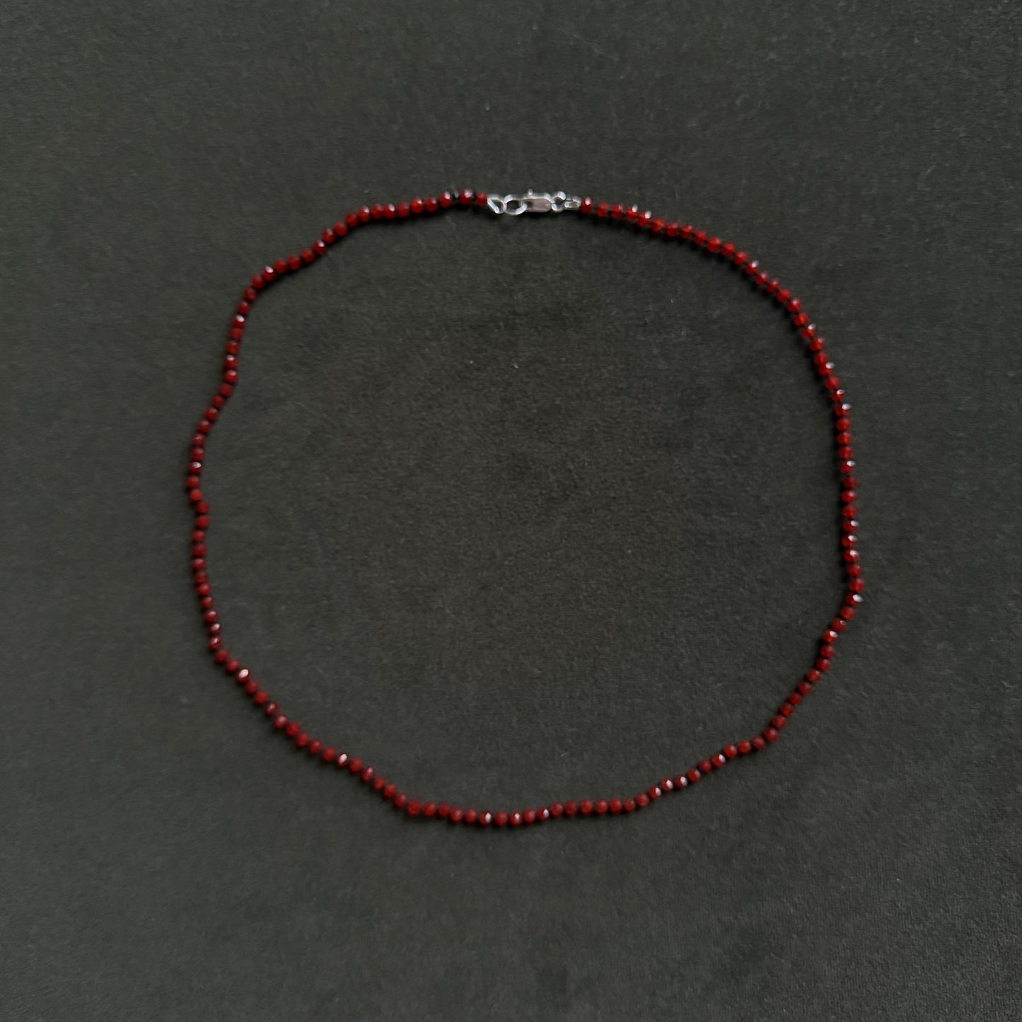 A beaded necklace made of small dark red faceted beads with a silver lobster clasp, displayed on a dark gray textured background.
