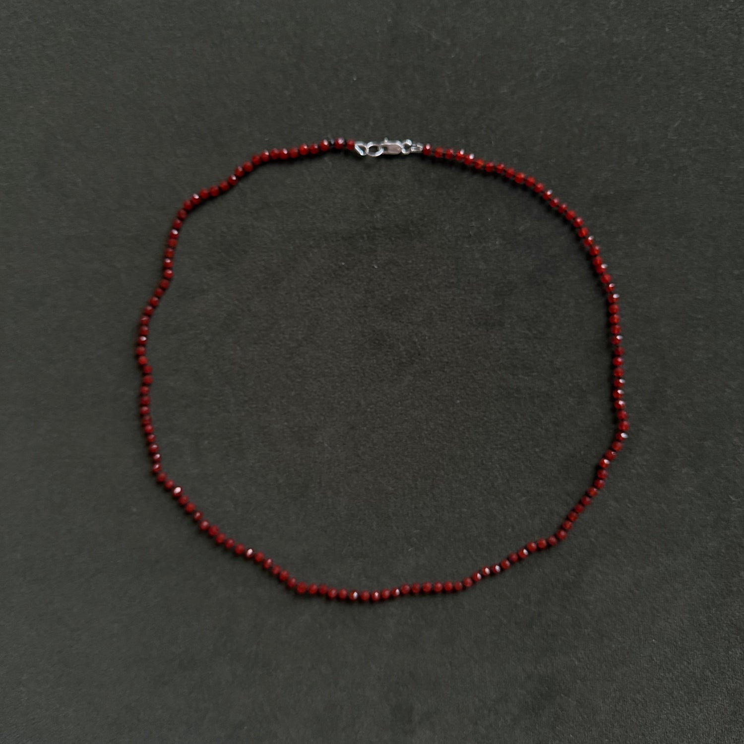 A beaded necklace made of small dark red faceted beads with a silver lobster clasp, displayed on a dark gray textured background.