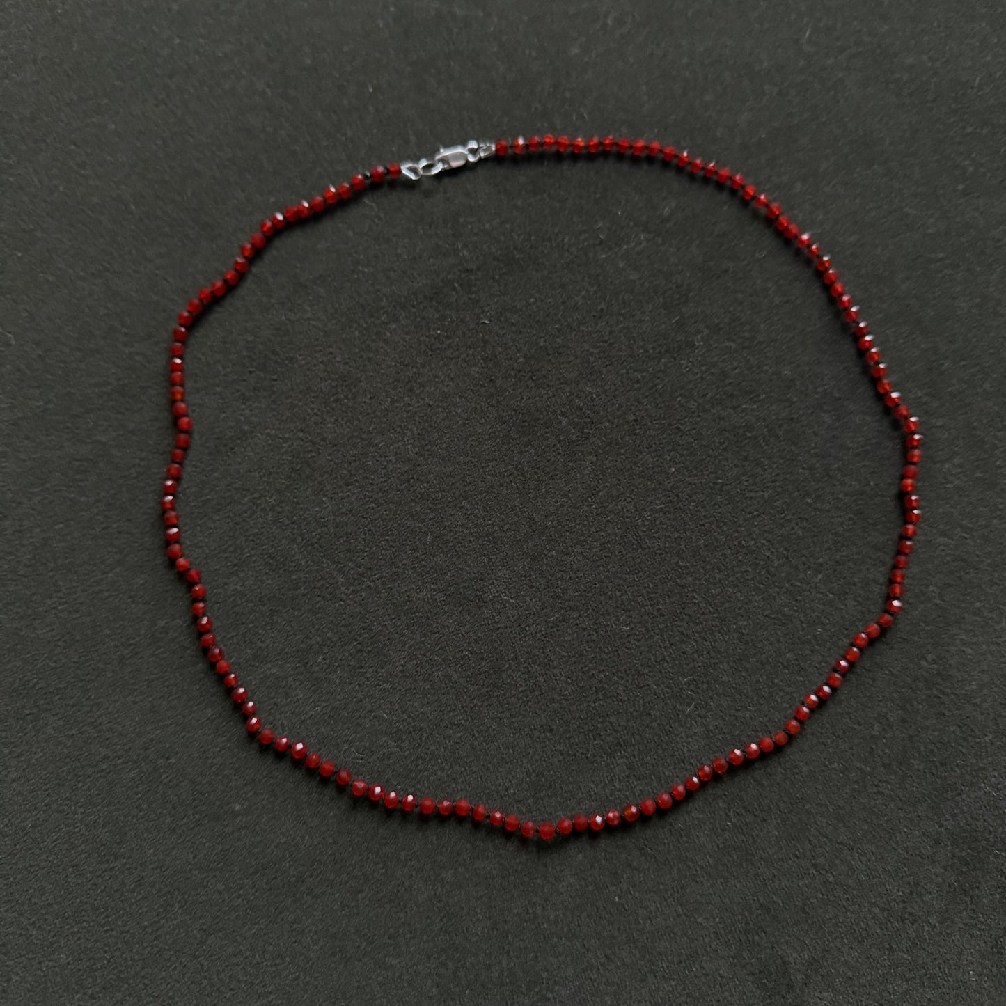 A beaded necklace made of small dark red beads with a silver lobster clasp, displayed on a dark gray textured background.