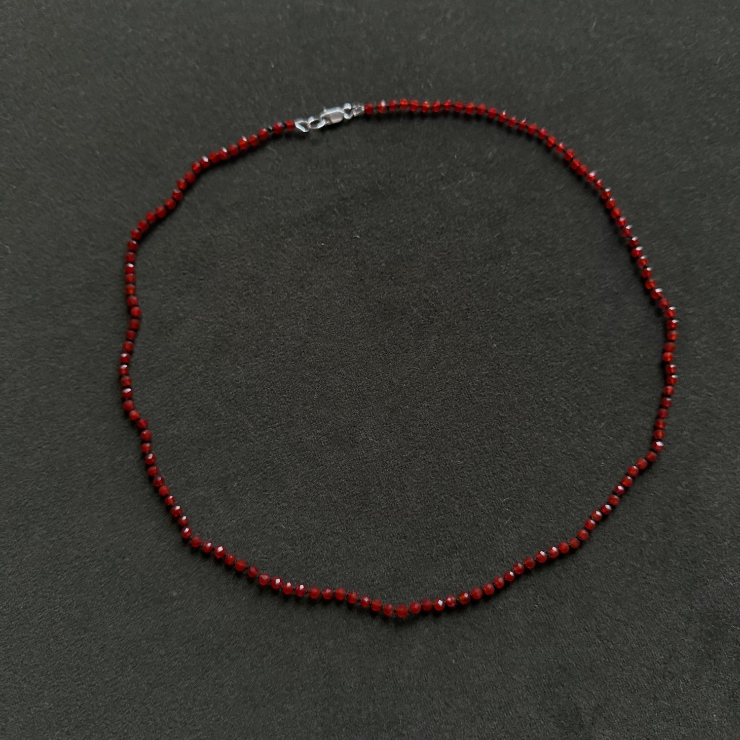 A beaded necklace made of small dark red beads with a silver lobster clasp, displayed on a dark gray textured background.