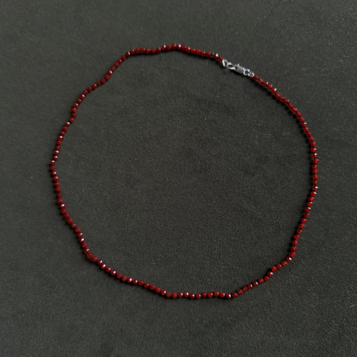 A beaded necklace made of small dark red faceted beads with a silver lobster clasp, laid flat on a dark gray textured background.