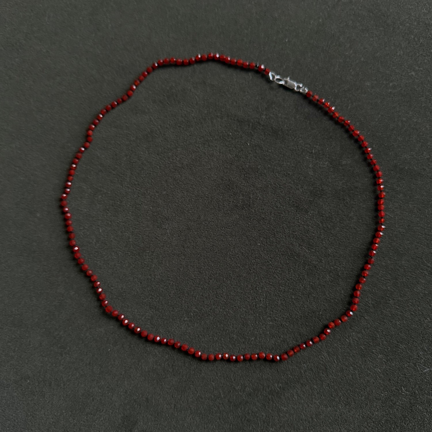 A beaded necklace made of small dark red faceted beads with a silver lobster clasp, laid flat on a dark gray textured background.