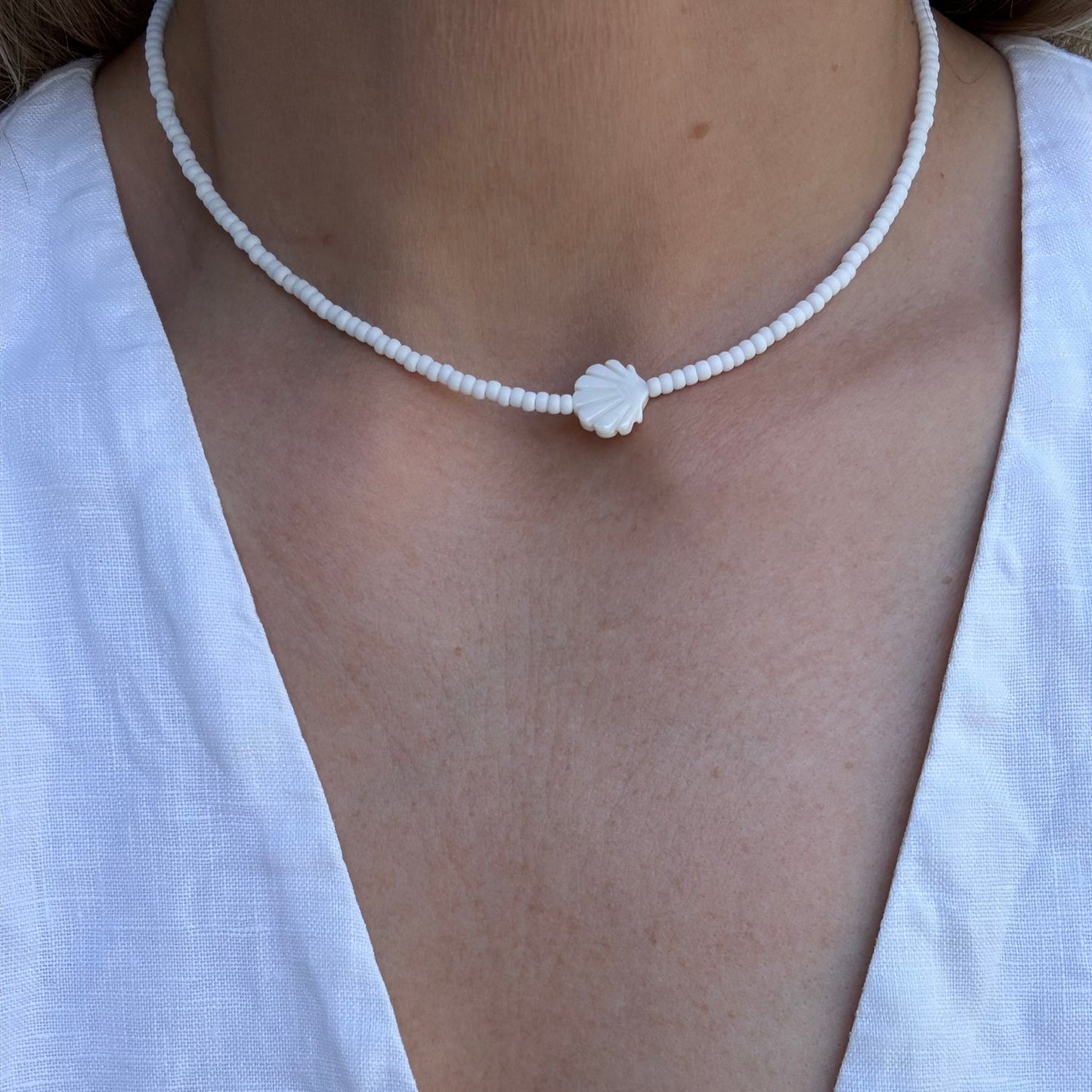 Person wearing a delicate white beaded necklace with a small round pendant on a plain background.