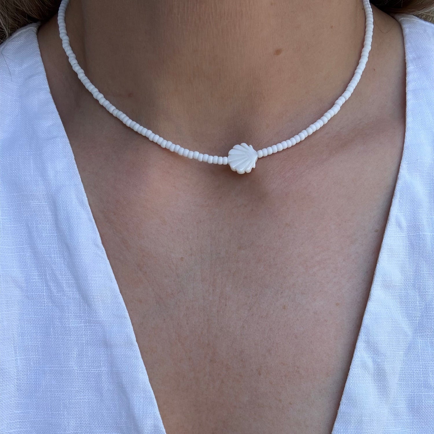 Person wearing a delicate white beaded necklace with a small round pendant on a plain background.