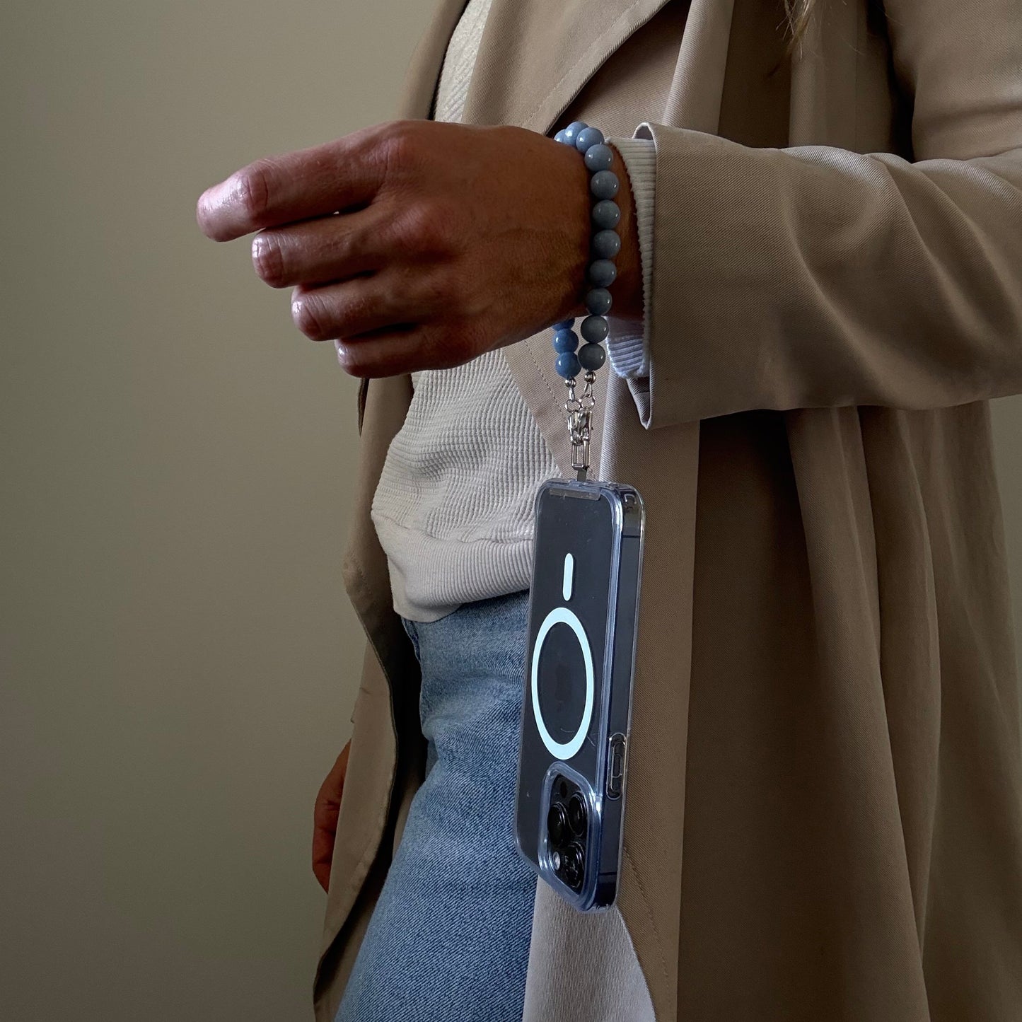 Person wearing a beige trench coat with a blue beaded mobile phone chain on a plain background