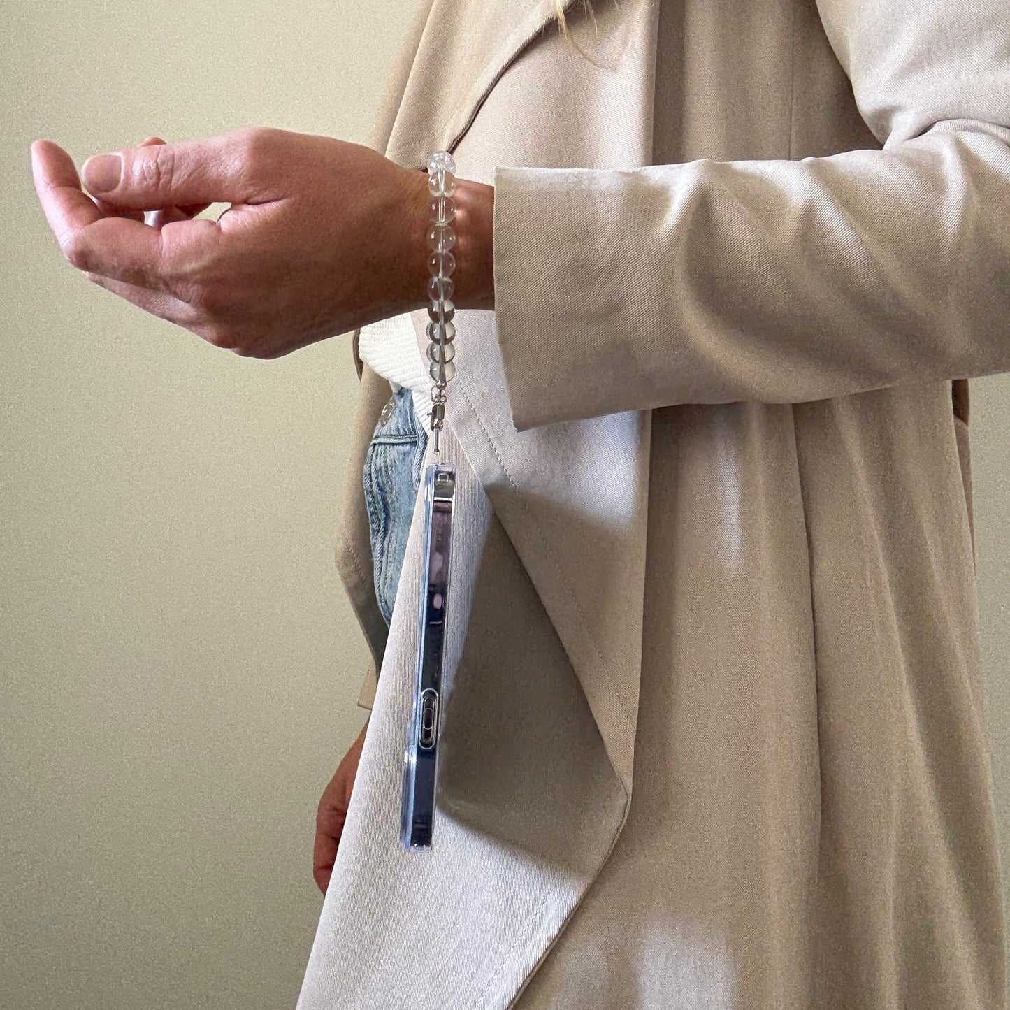 Person wearing a beige trench coat and blue jeans, holding a clear quartz crystal beaded phone chain.