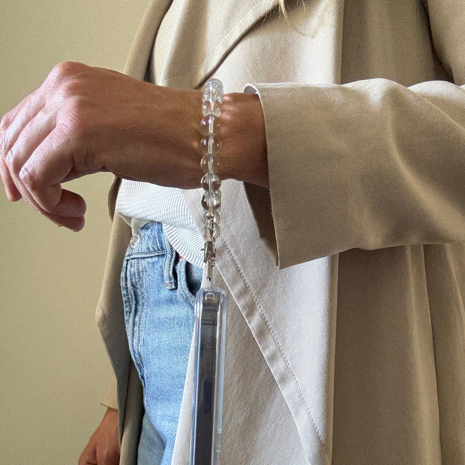 Person wearing a beige coat with a clear quartz crystal phone chain on a beige surface.