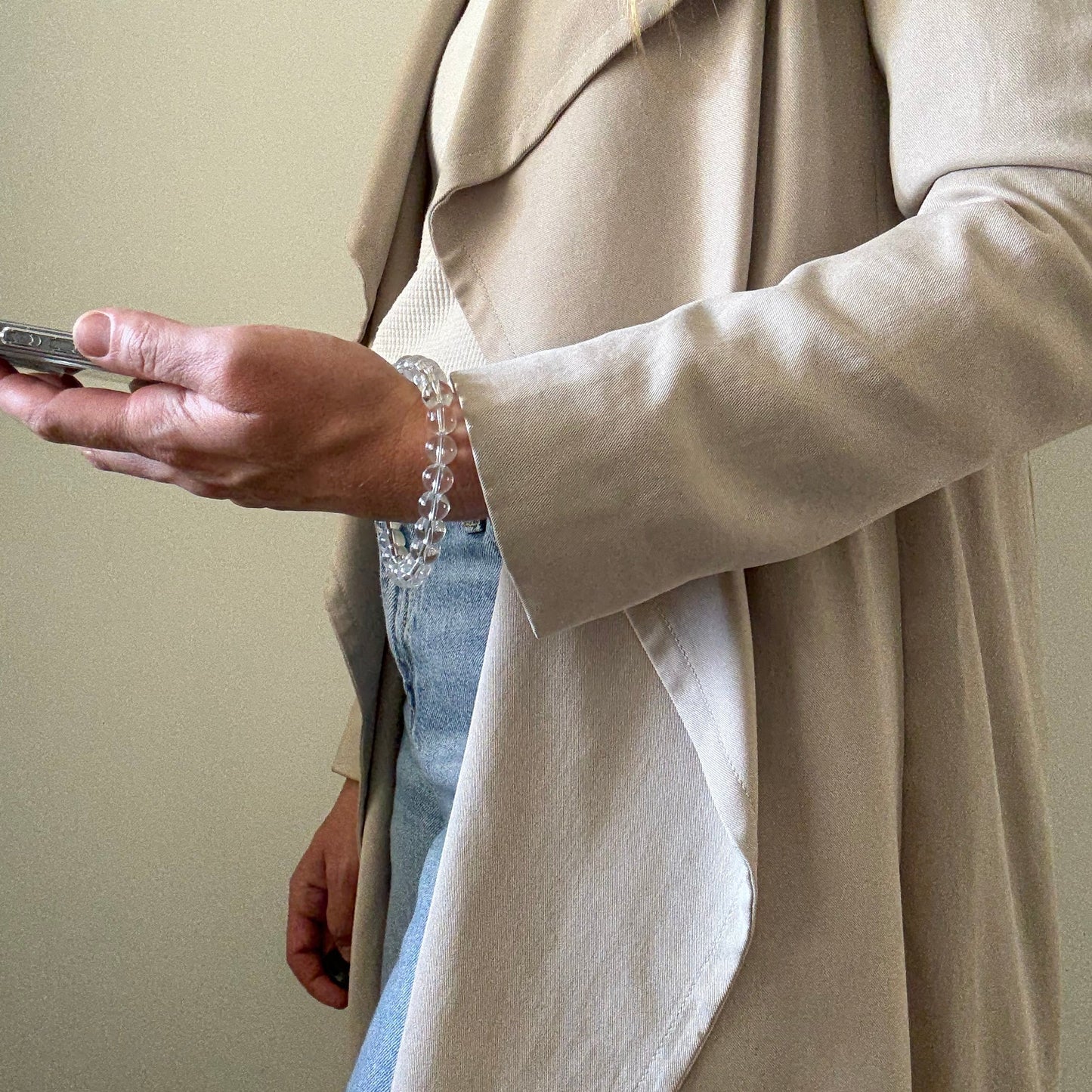 Person wearing a beige trench coat and blue jeans, holding a clear quartz crystal beaded phone chain.