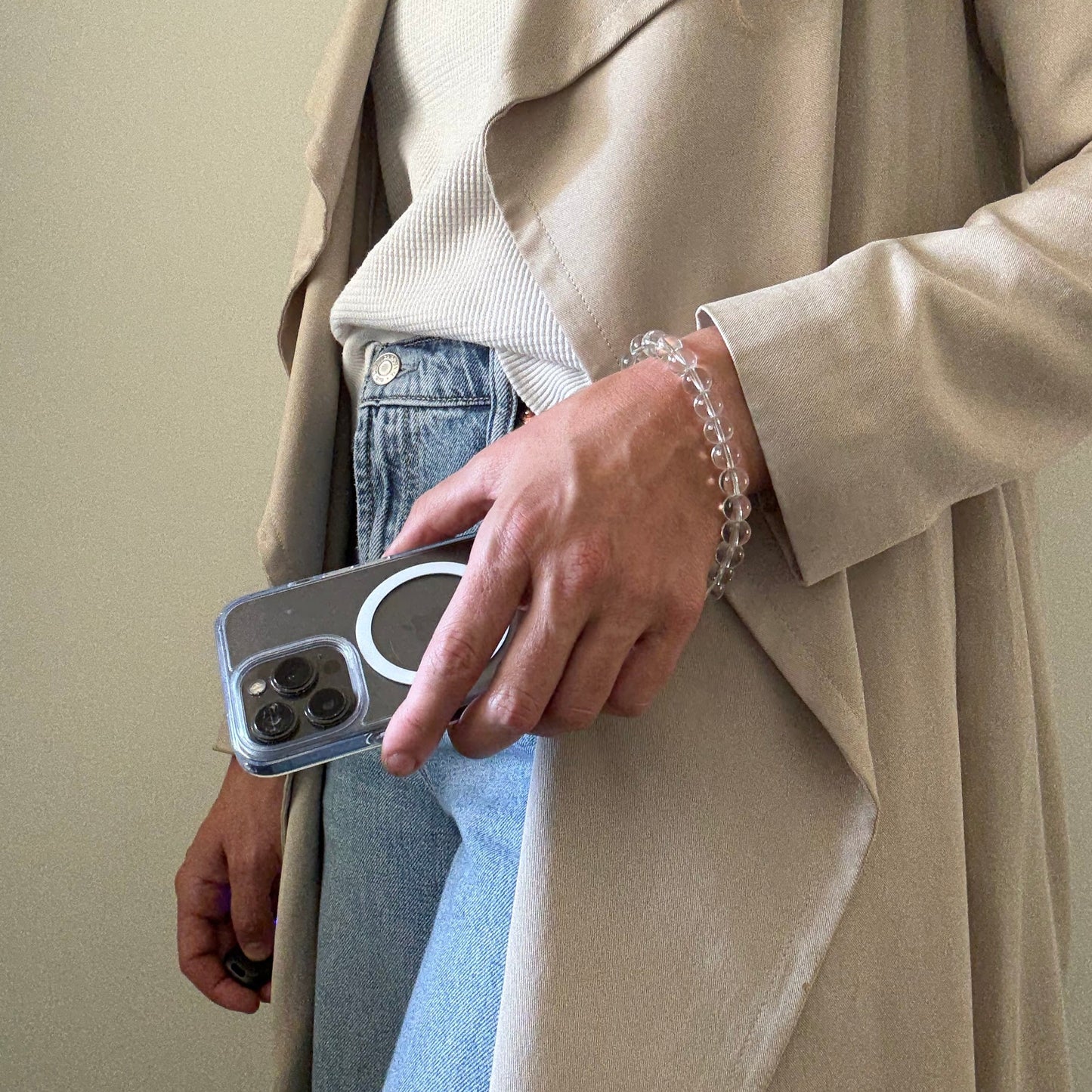Person wearing a beige trench coat and blue jeans, holding a clear quartz crystal beaded phone chain.