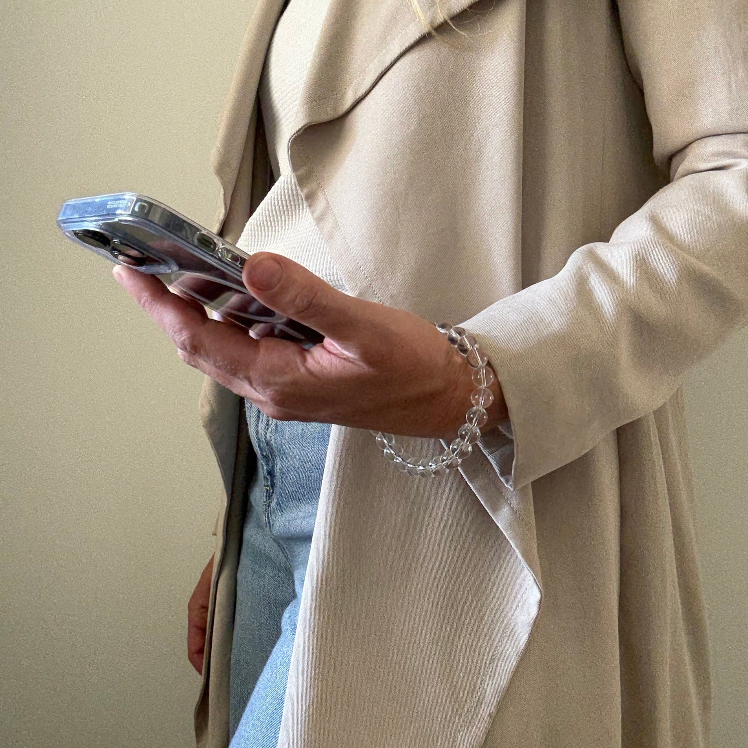 Person wearing a beige trench coat and blue jeans, holding a clear quartz crystal beaded phone chain.