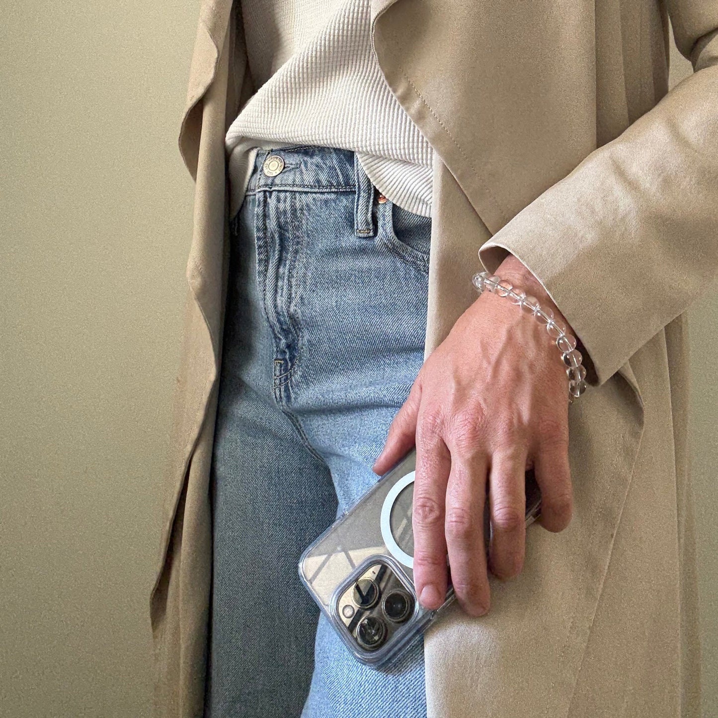 Person wearing a beige trench coat and blue jeans, holding a clear quartz crystal beaded phone chain.