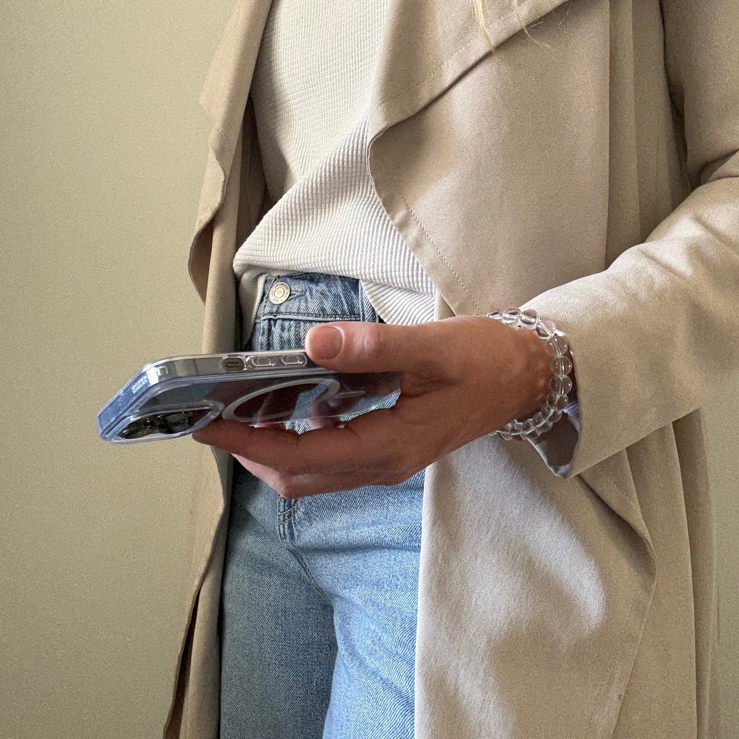 Person wearing a beige trench coat and blue jeans, holding a clear quartz crystal beaded phone chain.