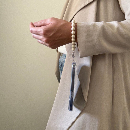 Close-up of a person wearing a beige trench coat with a hand holding a beaded fossil jasper phone chain