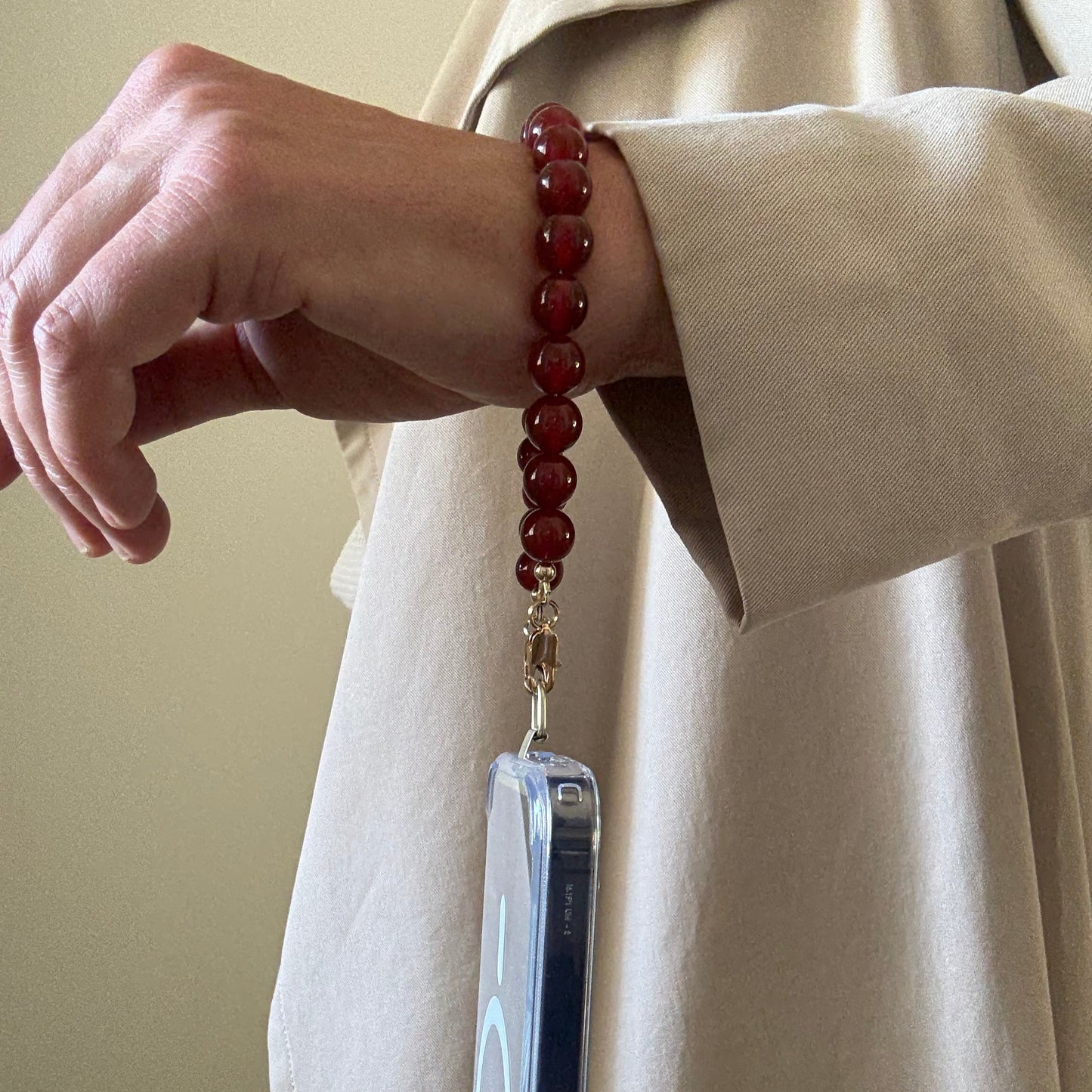 Hand holding a clear phone case with a red beaded phone strap against a beige background